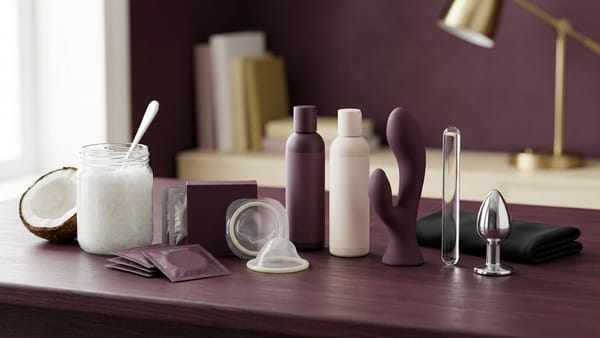 Dark plum desk with a coconut oil jar, coconut half, condom packets and barrier rings, lubricant bottles, and silicone, glass and stainless-steel sex toys.